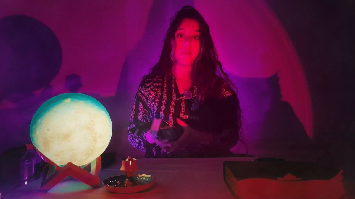 The image features a woman sitting at a table with a lamp, holding a deck of tarot cards. The woman appears to be engaged in a tarot reading session, possibly seeking guidance or insight into her life. A book can also be seen on the table, which might contain additional tarot card interpretations or other information related to tarot readings.