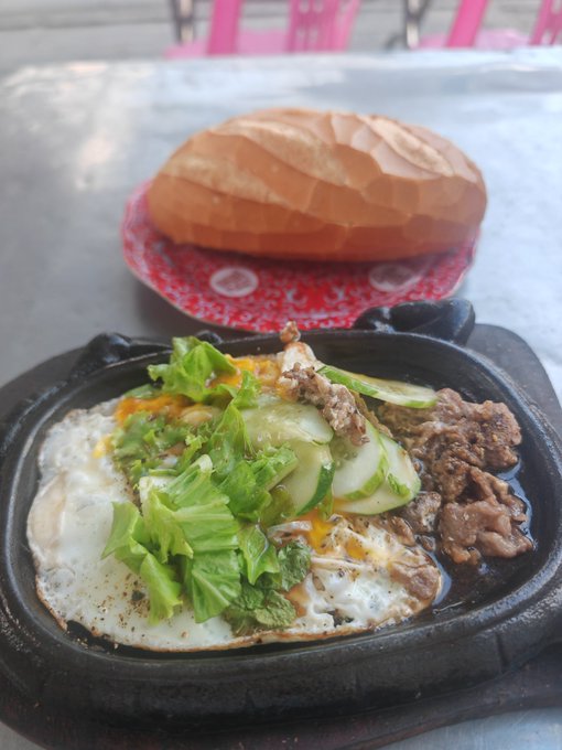 A Vietnamese dish featuring a sizzling cast iron skillet with beef, egg, and cucumber, accompanied by a crusty baguette on a red and white plate.