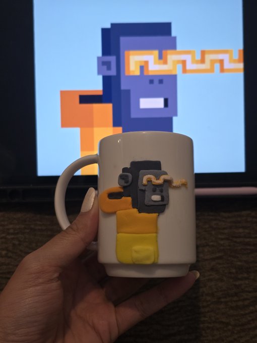 A hand holds a white ceramic mug decorated with a 3D clay figure resembling a pixelated character, with the same character displayed on a screen in the background.