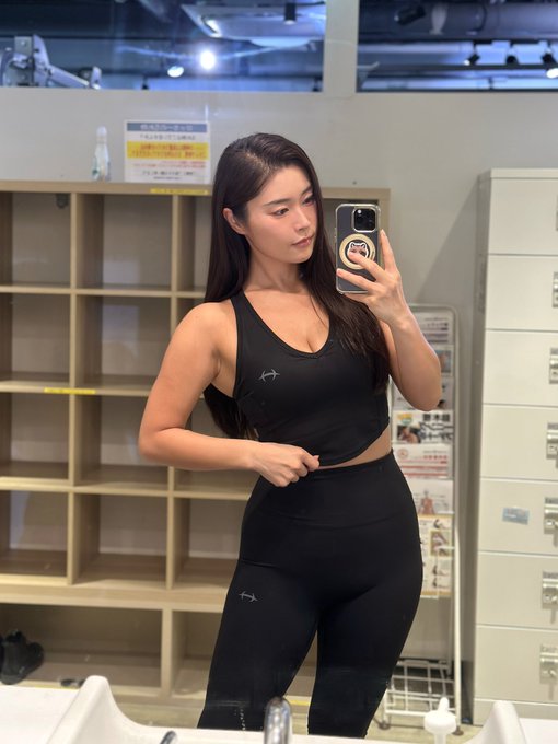 A fit young woman takes a mirror selfie at the gym, wearing a black athletic tank top and leggings, with lockers and cubbies visible in the background.