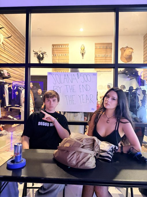 Two young people sit at a table holding up a handwritten sign reading "SOLANA $500 BY THE END THE YEAR," referencing the bold Solana price prediction tied to the "Solana Summerhouse" event. A backpack, drink can, and western-themed decor with animal skulls complete the scene.