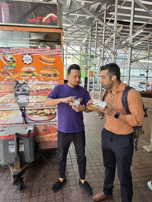 Two men stand in front of a New York City food cart advertising breakfast and lunch options, sharing foil-wrapped food.
