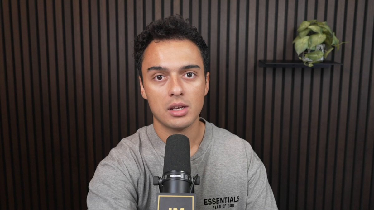 The image features a man wearing a gray shirt sitting behind a microphone with his hands on his lap. The background of the image shows a brown wall, which appears to be made of wood paneling. There are two potted plants visible in the scene - one located near the top left corner and another towards the right side of the image.