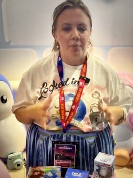 A woman wearing a graphic t-shirt and lanyard stands behind a display of various products and merchandise.