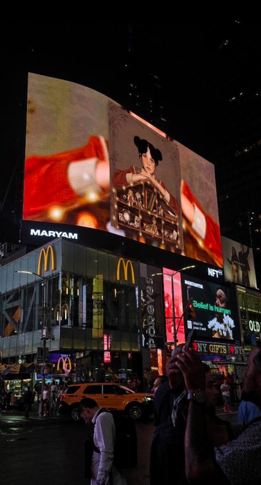 A nighttime cityscape shows a large digital billboard displaying a person with the text 'MARYAM' above a McDonald's and an 'NYC Gifts' shop, with people on the street below.