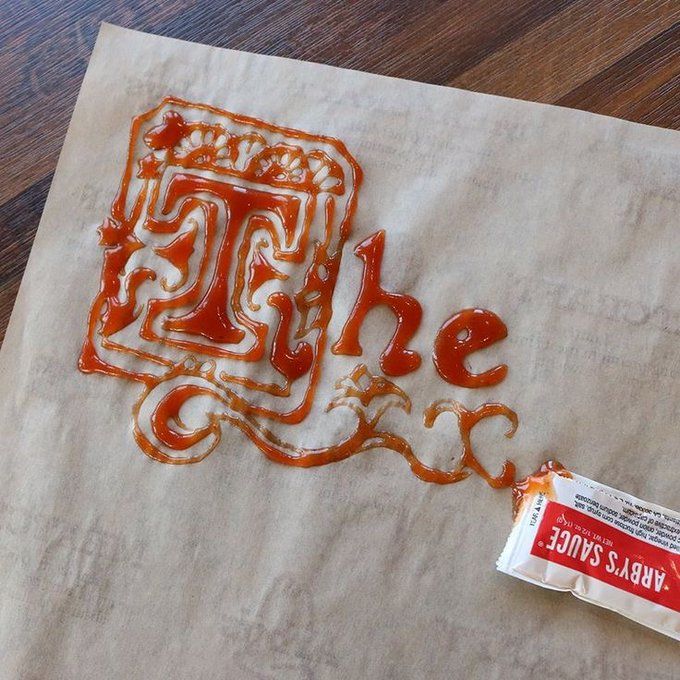 Red ketchup is squeezed from an 'ARBY'S SAUCE' packet onto parchment paper, artistically spelling out 'The' and decorating a large letter 'T'.