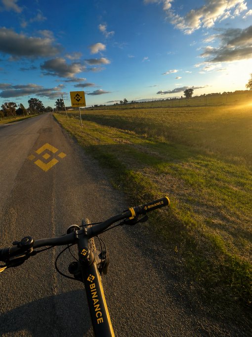 A first-person view from a yellow Binance-branded bicycle handlebar captures a peaceful countryside road at golden hour, complete with a matching Binance-branded trail marker sign down the path—fitting perfectly with a casual post simply saying "Just out for a ride."