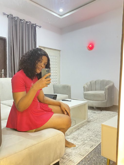 A mirror selfie of a woman wearing a red dress, seated on a light-colored couch in a modern living room with a pink light on the wall.