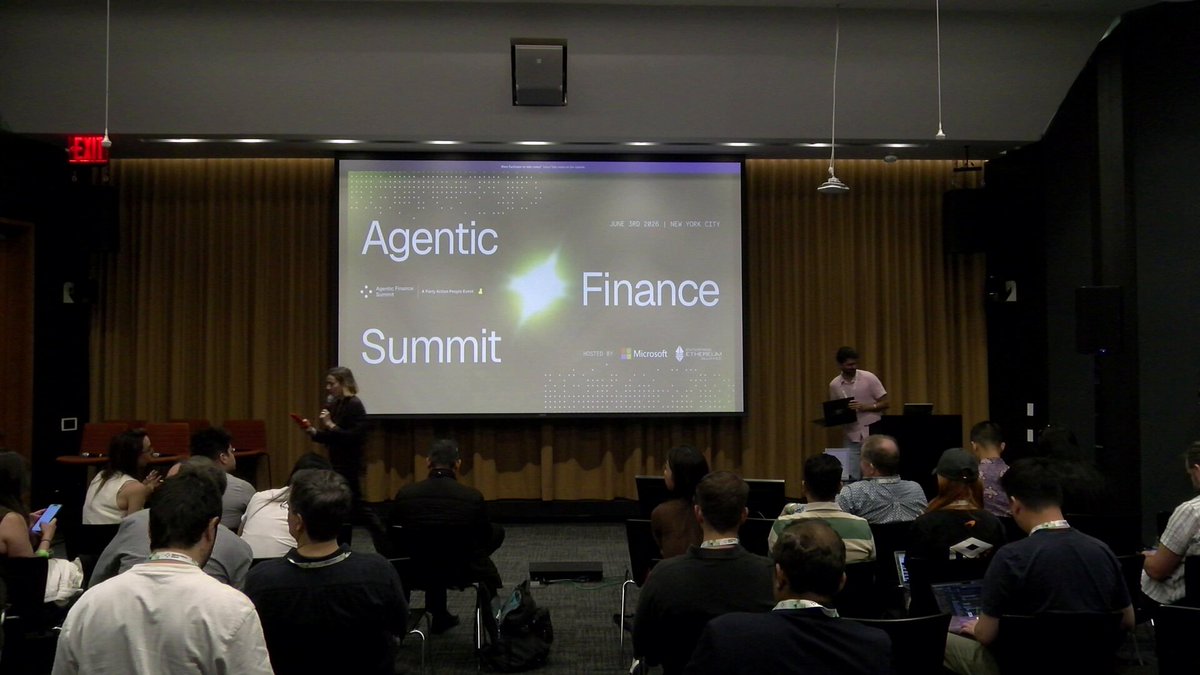 In this image, a group of people are gathered around a large screen displaying information about an event called "Agents need wallets, execution, and payment rails that work together." The event appears to be a summit or conference related to agentic finance. There is a speaker on stage addressing the audience, who seem engaged in the presentation.

The room has multiple chairs arranged around the space, with some people sitting down while others are standing. A few individuals can be seen holding their cell phones, possibly taking notes or capturing photos of the event. The overall atmosphere suggests that this is a formal gathering focused on discussing and learning about agentic finance.