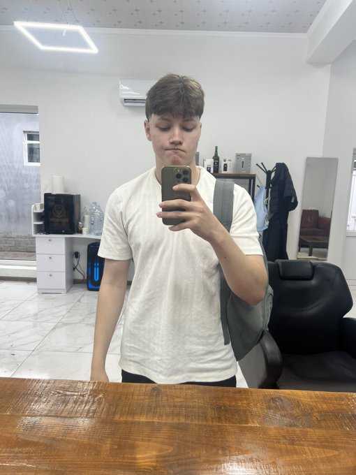 Mirror selfie of a young man in a white t-shirt taking a photo with his phone in what appears to be a salon or barbershop.