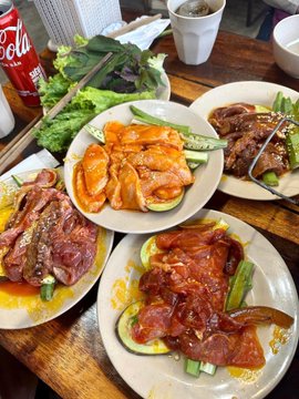 An overhead view of a meal featuring plates of Korean-style spicy marinated meat, cucumber slices, fresh greens, and a can of Coca-Cola on a wooden table.