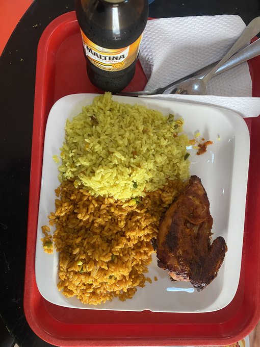 A red tray holds a meal consisting of yellow rice, seasoned grain, and a piece of roasted meat, accompanied by a bottle of "MARTINA" beer.