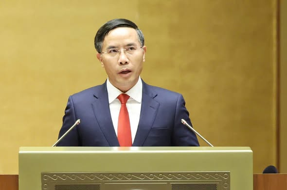 A man in a dark suit and red tie speaking at a golden podium with two microphones, against a plain yellow background.