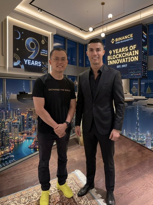 Two men pose together in front of a "Binance 9 Years" and "9 Years of Blockchain Innovation - Est. 2017" display, with one wearing a black "Exchange The World" t-shirt and bright yellow sneakers, while the other is dressed in a black suit, celebrating Binance's 9th anniversary milestone.