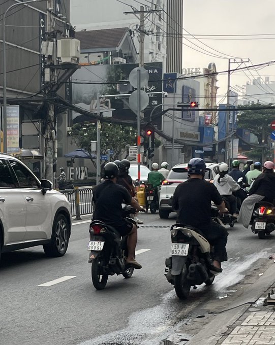 A street scene in Vietnam with multiple motorcyclists riding on a wet road, alongside a white car, with tangled overhead power lines and storefronts lining the street.
