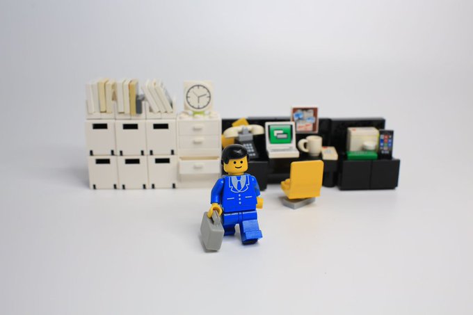 A LEGO figurine of a businessman in a blue suit carrying a briefcase, posed in front of a miniature office setup with bookshelves, a desk, computer, and clock.
