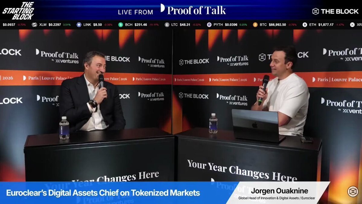 The image shows two men sitting at a table with microphones, appearing to be discussing something related to digital assets or cryptocurrencies. The background features a large banner displaying various logos of companies such as Euroclear, Proof of Talk, and others. The text on the banner suggests that these companies are involved in managing digital assets and have significant financial resources.