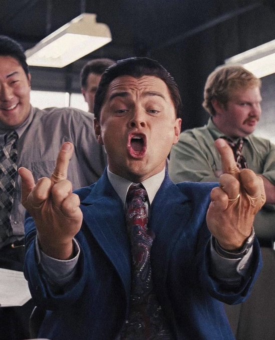 A still from a film featuring Leonardo DiCaprio in a blue pinstripe suit making an exaggerated facial expression with both middle fingers raised.
