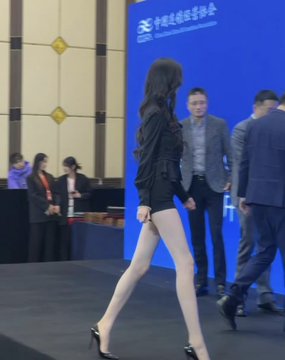 The image features a woman wearing black clothing, including short shorts, walking down a runway with her legs crossed. She is positioned in front of a blue wall that has Chinese characters written on it. The text "This leg is truly so authoritative." appears below the woman's feet, suggesting that she might be the center of attention at an event or gathering.

There are several people present in the background, including one person standing behind the woman and another person walking towards her from the right side of the image. A few more individuals can also be seen scattered around the scene, indicating a social setting where this event is taking place.
