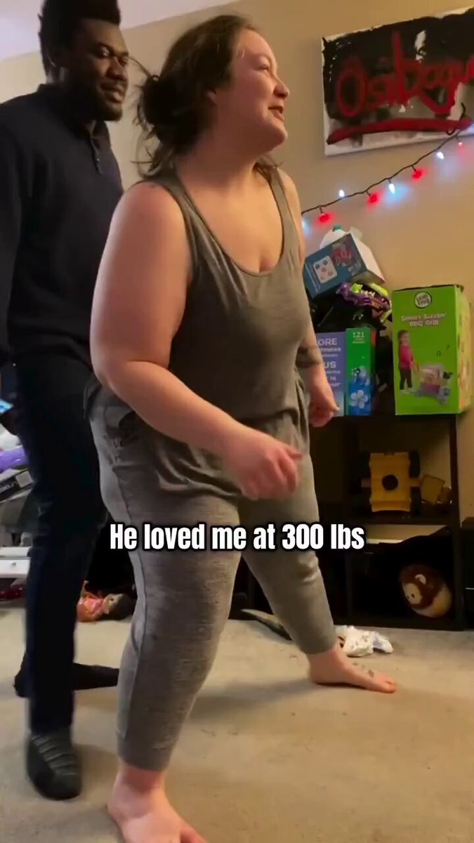 The image shows a man standing next to a woman who appears to be dancing, possibly wearing workout clothes. They are both barefoot, which adds to the casual atmosphere of the scene. The background features a wall with various items such as books and toys, suggesting that they might be in a living room or playroom setting. A text overlay on the image reads "He loved me at 300 lbs". This message is likely meant to convey the man's appreciation for the woman's weight loss journey, possibly related to their shared interest in fitness or health.