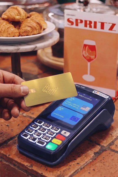 A hand holds a gold Ether.fi card over a payment terminal displaying a currency selection screen with "Local Currency" and USD options, next to a Spritz menu and croissants at a café. The image illustrates the post's message about choosing local currency at checkout to avoid the terminal's 5-6% FX markup and instead benefit from Ether.fi's 0% FX rate.