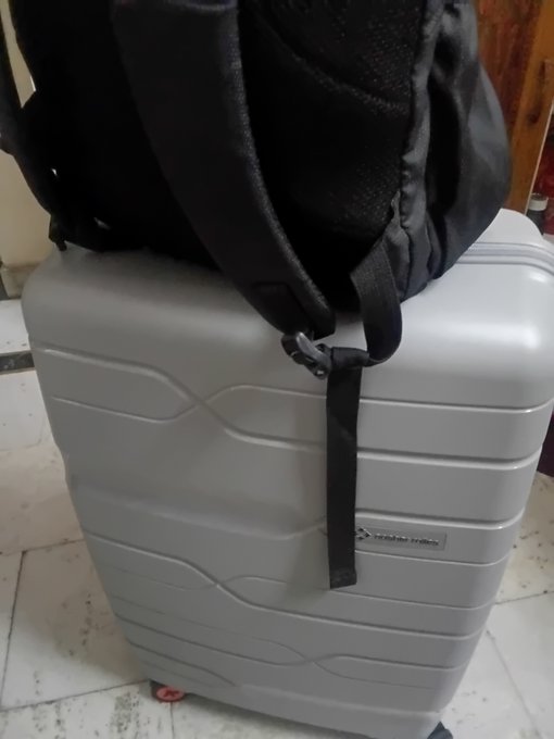 A silver suitcase with a black backpack strapped to the top sits on a tiled floor.