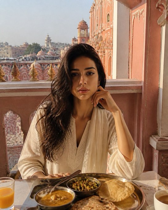 A woman with long dark hair sits at a table eating a meal, with a stunning view of ornate, reddish-pink buildings and temples in the background.