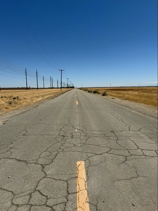 A long, cracked asphalt road stretches into the distance through a flat, dry landscape under a clear blue sky, lined with utility poles.