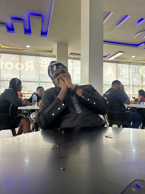 A man in traditional black attire and patterned cap sits at a black table with his hands folded near his face, inside what looks like a food court with other patrons in the background.