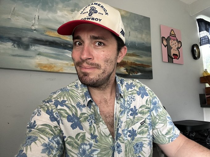 A man wearing a 'COWBOY' cap and a blue and green floral Hawaiian shirt poses in front of a painting, with a pixel-art monkey artwork visible on the wall behind him.