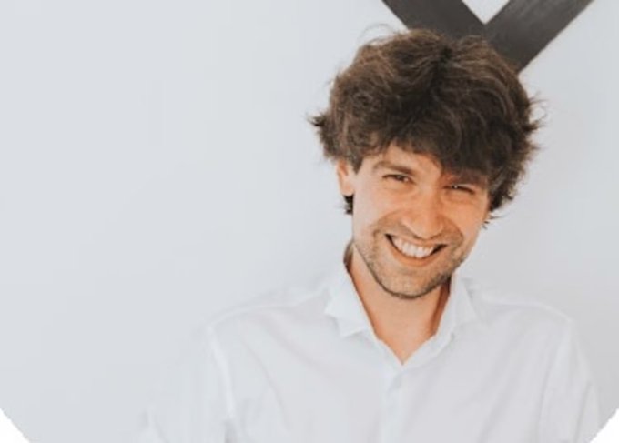 The image features a man wearing a white shirt, smiling at the camera with an expression of happiness or amusement. The background consists of a plain white wall, which contrasts with the man's attire and adds depth to the scene. There are no texts or other objects in the image, making it a simple yet engaging composition that captures the viewer's attention.