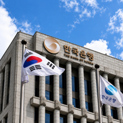 The image shows the Bank of Korea headquarters building, identifiable by its name displayed in both Korean (한국은행) and English, along with the South Korean national flag flying in the foreground, directly relating to the accompanying article about the Bank of Korea's study on how dollar-backed stablecoins like USDT and USDC can drive down local currencies like the Korean won.