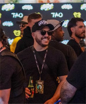 A man wearing a black cap and sunglasses smiling while holding a beer at a venue with 'TAO' logos visible in the background.
