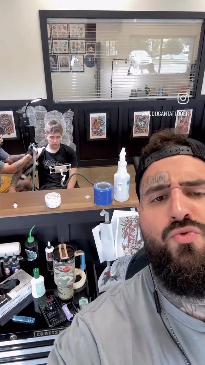 A selfie video screenshot of a bearded tattoo artist (@WUDUGANTATTOOER) in a tattoo studio with a young client sitting in the chair behind him.