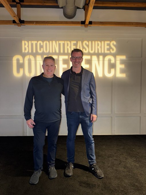 Two men stand smiling in front of a wall with the projected text 'BITCOINTREASURIES CONFERENCE'.