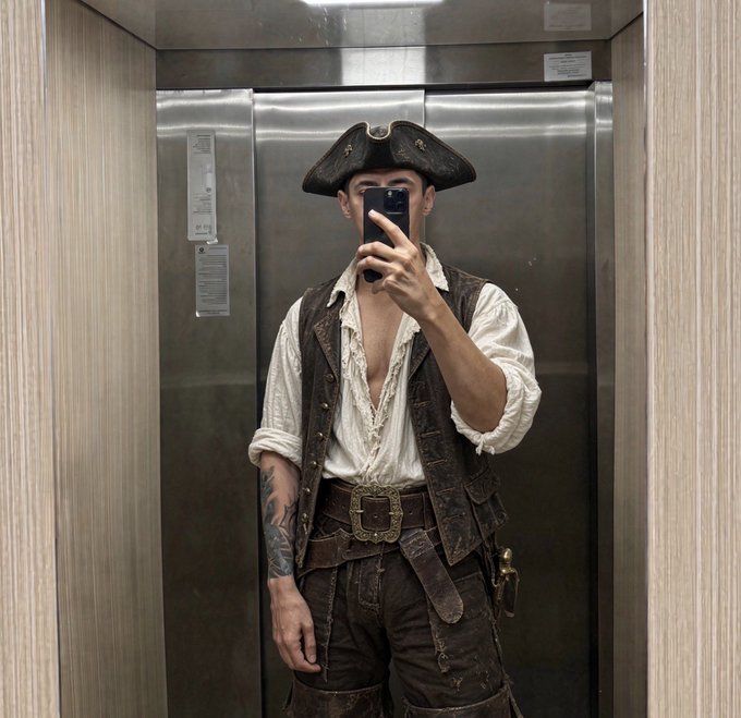 A mirror selfie of a person dressed in a pirate costume featuring a tricorn hat, a leather vest over an open white shirt, and a wide ornate belt, taken inside an elevator.
