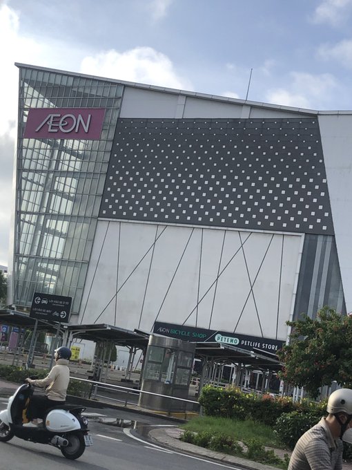 An exterior photograph of a large Aeon shopping mall featuring a modern black and white facade, with a person riding a white scooter on the street in the foreground.