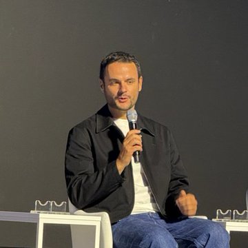 Johann Kerbrat, a representative from Robinhood, speaks into a microphone while seated on stage at Korea Blockchain Week 2026, likely addressing the company's stock token custody practices. He wears a black jacket over a light shirt, seated against a dark backdrop in what appears to be a panel discussion setting.