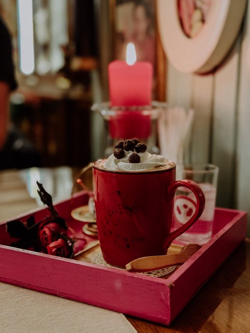 A cozy café scene featuring a red mug topped with whipped cream and chocolate-covered coffee beans, displayed on a red tray alongside a lit pink candle and dried roses.