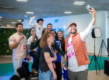 A diverse group of enthusiastic volunteers smile and pose energetically for a selfie at an indoor event space, capturing the lively community spirit needed to help bring ETHGlobal Lisbon & Pragma to life this July 24-26. The joyful expressions and team camaraderie in the image embody the welcoming, behind-the-scenes vibe the post is seeking to promote.
