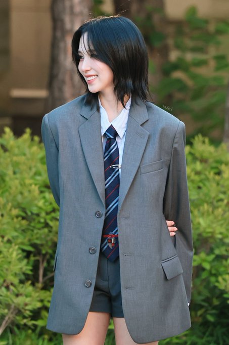 A smiling young woman with shoulder-length dark hair poses outdoors in an oversized grey blazer, white shirt, and striped tie.