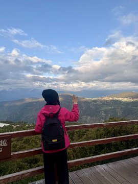 Photo of a person wearing a pink jacket, black beanie, and backpack standing at a wooden railing overlooking a vast mountain range under a partly cloudy sky.