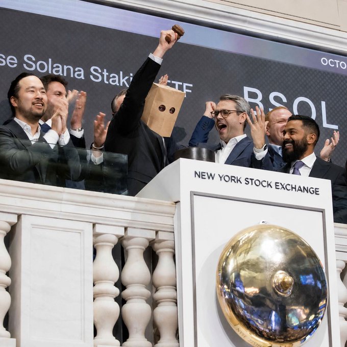 A celebratory scene at the New York Stock Exchange with a group of men in suits cheering, one wearing a paper bag over his head with drawn-on eyes, as the Solana Staking ETF begins trading.
