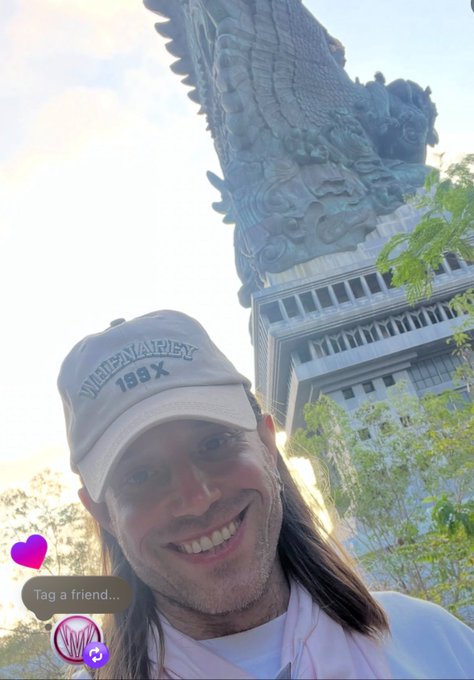 A selfie of a smiling person with long hair wearing a "Wheewarey 199X" cap, posing in front of an ornate temple-like structure with a dragon-shaped sculpture on top, with a "Tag a friend..." sticker visible on the screen. The image likely relates to Jared, the CEO of an NFT platform, showing him enjoying a scenic moment at what appears to be a cultural landmark.