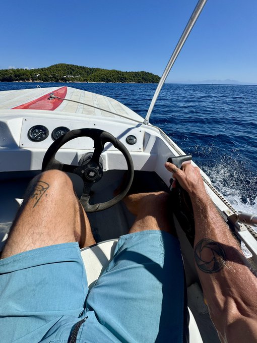 A first-person view of someone driving a boat on blue water toward a forested island, with tattoos visible on their arms.