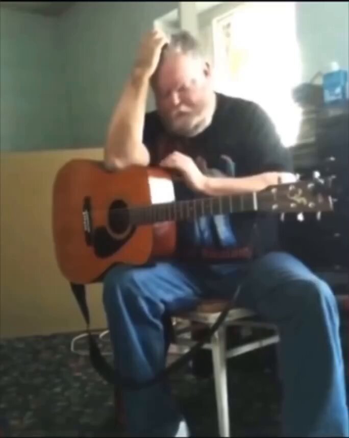 The image features an older man sitting down with a guitar, holding his head up as if he's scratching or rubbing his head. He appears to be wearing a black shirt and blue jeans. The background of the photo shows a chair where the man is seated, and there are several books scattered around him. The caption for this image reads: "$pork".