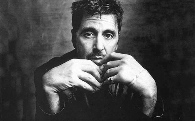 A black and white portrait of a man with dark hair and a beard, resting his chin on his clasped hands, with visible tattoos on his forearm.
