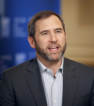A man with a short beard wearing a dark suit jacket over a light blue checkered shirt, speaking with his mouth open against a blue background.