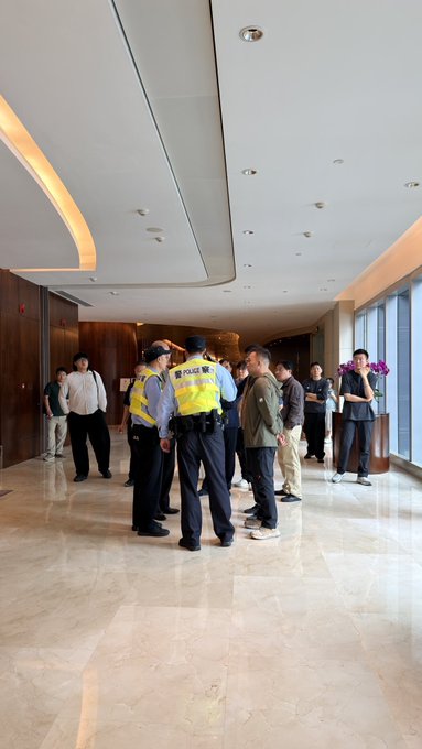 Two police officers in yellow vests marked "POLICE 警察" converse with a group of men in a polished lobby, suggesting a public scene where quiet financial ambition and visible authority intersect.