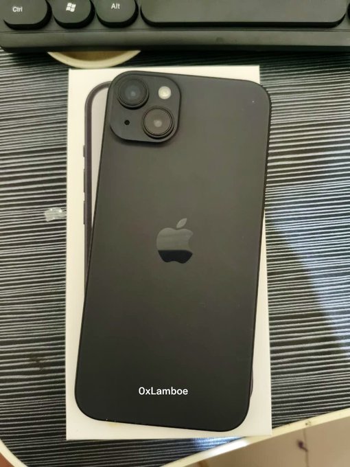 A black iPhone (likely a 14 or 15 model based on the dual-camera layout) rests face-down on a white surface atop a dark textured mat, with a keyboard partially visible in the background; the photo is watermarked '0xLamboe'.